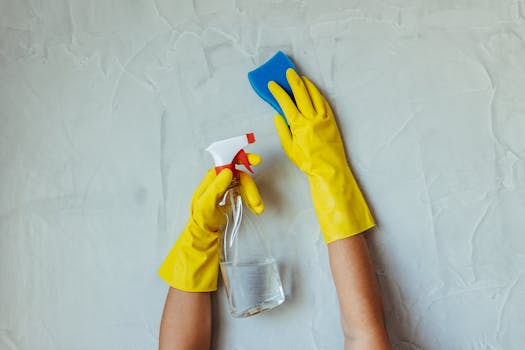 Sodium Bicar Close-up of hands in yellow gloves cleaning a wall with a spray bottle and sponge.