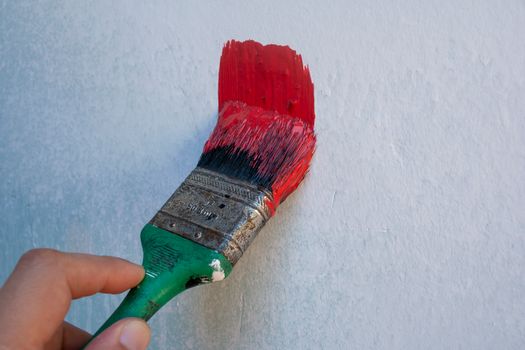 Detailed image of a hand holding a paintbrush with red paint on a textured wall.