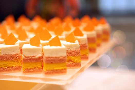 Row of neatly arranged colorful layered cakes with orange triangular toppings on a white platter.