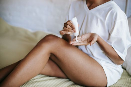 Unrecognizable female in white t shirt with bared legs sitting on bed and applying cream on hand while doing skincare routine